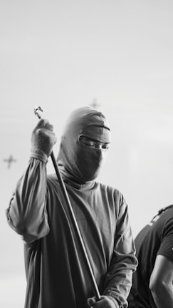 A person wearing a full-body suit with a face cover, holding a long pole. The suit is reminiscent of protective or specialized workwear. The setting appears to be indoor with a plain background, focusing on the individual and their attire.