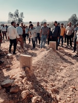 A group of people standing around two fresh mounds of earth in an outdoor setting, likely a burial site. The scene includes men dressed in casual clothing, suggesting a communal or commemorative gathering. The area is surrounded by dry terrain with scattered rocks and some trees in the background.