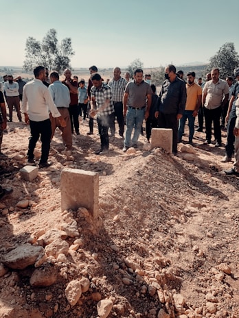 A group of people standing around two fresh mounds of earth in an outdoor setting, likely a burial site. The scene includes men dressed in casual clothing, suggesting a communal or commemorative gathering. The area is surrounded by dry terrain with scattered rocks and some trees in the background.