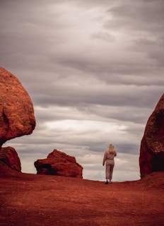 A person walks between large, reddish rock formations under a cloudy sky, creating a sense of solitude and exploration.