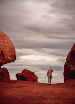 A person walks between large, reddish rock formations under a cloudy sky, creating a sense of solitude and exploration.