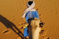 a man in a blue robe walking a camel in the desert