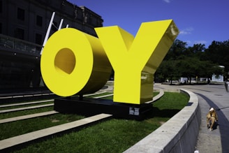 Large yellow letters spelling 'OY' are positioned on a black platform in an outdoor park setting. The bright color of the letters stands out against the surrounding green grass and trees. A dog on a leash walks along the sidewalk, with part of the person holding the leash unseen. In the background, there is a modern building with a glass facade.