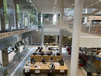 A modern library interior with an open and spacious layout. Large glass windows provide ample natural light. People are seated at long wooden tables, some engaging with electronic devices and books. The space has multiple levels with stairs leading up to an upper floor, and there are signs indicating different areas. The surroundings are clean and organized.
