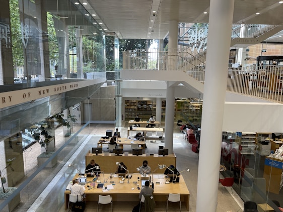 A modern library interior with an open and spacious layout. Large glass windows provide ample natural light. People are seated at long wooden tables, some engaging with electronic devices and books. The space has multiple levels with stairs leading up to an upper floor, and there are signs indicating different areas. The surroundings are clean and organized.