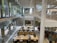 A modern library interior with an open and spacious layout. Large glass windows provide ample natural light. People are seated at long wooden tables, some engaging with electronic devices and books. The space has multiple levels with stairs leading up to an upper floor, and there are signs indicating different areas. The surroundings are clean and organized.