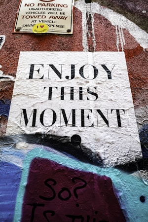 A textured wall featuring various street art elements with a prominent white paper sign displaying the phrase 'ENJOY THIS MOMENT' in bold black letters. Above it is a weathered 'NO PARKING' sign with a small yellow sticker that has a heart symbol and text. The background includes abstract graffiti with layered colors such as purple, blue, and black.