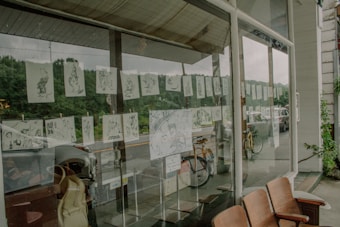 A series of illustrated sketches are displayed on a large window, facing an outdoor street scene with parked bicycles and cars. The illustrations are suspended in rows and appear to be black and white, depicting various scenes or characters. Reflections in the glass show greenery and a cloudy sky.