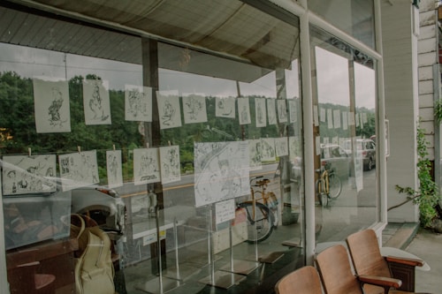 A series of illustrated sketches are displayed on a large window, facing an outdoor street scene with parked bicycles and cars. The illustrations are suspended in rows and appear to be black and white, depicting various scenes or characters. Reflections in the glass show greenery and a cloudy sky.