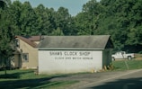 A small building labeled 'Shaws Clock Shop' with services for clock and watch repair is situated by a road. The structure is a single-story block with a pitched roof, adjacent to a larger beige house with a brown roof. Lush greenery and tall trees surround the area, and a white pickup truck is parked on the grass.