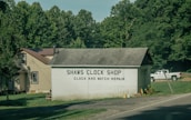 A small building labeled 'Shaws Clock Shop' with services for clock and watch repair is situated by a road. The structure is a single-story block with a pitched roof, adjacent to a larger beige house with a brown roof. Lush greenery and tall trees surround the area, and a white pickup truck is parked on the grass.