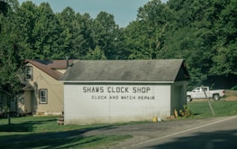 A small building labeled 'Shaws Clock Shop' with services for clock and watch repair is situated by a road. The structure is a single-story block with a pitched roof, adjacent to a larger beige house with a brown roof. Lush greenery and tall trees surround the area, and a white pickup truck is parked on the grass.