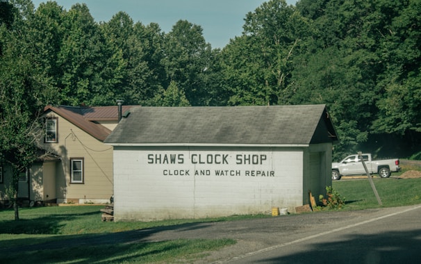 A small building labeled 'Shaws Clock Shop' with services for clock and watch repair is situated by a road. The structure is a single-story block with a pitched roof, adjacent to a larger beige house with a brown roof. Lush greenery and tall trees surround the area, and a white pickup truck is parked on the grass.