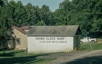 A small building labeled 'Shaws Clock Shop' with services for clock and watch repair is situated by a road. The structure is a single-story block with a pitched roof, adjacent to a larger beige house with a brown roof. Lush greenery and tall trees surround the area, and a white pickup truck is parked on the grass.