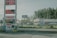 A roadside scene featuring a large sign displaying gas prices and a digitally lit 'Help Wanted' message. Below is a 'Liquor' sign accompanied by various advertisements for beer and snacks. The background includes a strip mall with a pet store and other businesses nestled among pine trees.