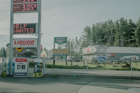 A roadside scene featuring a large sign displaying gas prices and a digitally lit 'Help Wanted' message. Below is a 'Liquor' sign accompanied by various advertisements for beer and snacks. The background includes a strip mall with a pet store and other businesses nestled among pine trees.