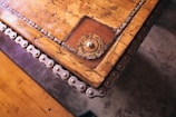 A wooden tabletop with intricate metal chains along the edges, featuring a gear-like object embedded in one corner. The surface is rustic with visible grains and scratches, enhancing its vintage appeal.