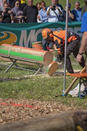 A person wearing orange protective gear and a helmet operates a chainsaw to cut through large logs. The event takes place outdoors with a crowd watching in the background. The logs are mounted on metal stands on a grassy area, and there is a visible banner advertising in the background.
