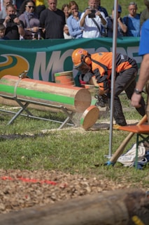 A person wearing orange protective gear and a helmet operates a chainsaw to cut through large logs. The event takes place outdoors with a crowd watching in the background. The logs are mounted on metal stands on a grassy area, and there is a visible banner advertising in the background.