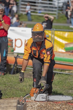 A person wearing protective gear such as a helmet, visor, and gloves is operating a chainsaw at an outdoor event. The background features a crowd behind protective barriers and a grassy area.