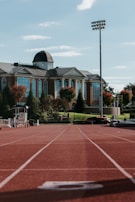 A running track leads to a large brick building with white columns and a dome on the roof. The building is surrounded by landscaped greenery and trees with autumn foliage. A tall light pole stands to the right, and various sports equipment is visible near the track.