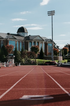 A running track leads to a large brick building with white columns and a dome on the roof. The building is surrounded by landscaped greenery and trees with autumn foliage. A tall light pole stands to the right, and various sports equipment is visible near the track.