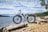 A white electric bicycle is positioned on a rocky shore near a body of water under a partly cloudy sky. The background includes a view of the water extending to the horizon with forested areas visible in the distance. There are some green trees on the right side.