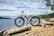 A white electric bicycle is positioned on a rocky shore near a body of water under a partly cloudy sky. The background includes a view of the water extending to the horizon with forested areas visible in the distance. There are some green trees on the right side.