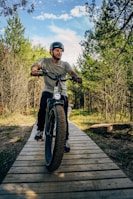 A person wearing a helmet rides a fat tire bike along a wooden path surrounded by lush, green trees under a partly cloudy sky. The scene conveys a sense of adventure and outdoor exploration.
