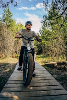 A person wearing a helmet rides a fat tire bike along a wooden path surrounded by lush, green trees under a partly cloudy sky. The scene conveys a sense of adventure and outdoor exploration.