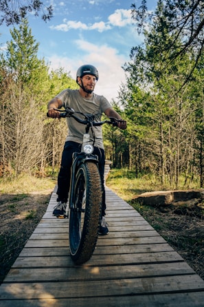 A person wearing a helmet rides a fat tire bike along a wooden path surrounded by lush, green trees under a partly cloudy sky. The scene conveys a sense of adventure and outdoor exploration.