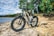 An electric bicycle is parked on rocky terrain by the edge of a body of water with lush greenery in the background. The sky is partly cloudy, suggesting a bright, pleasant day.