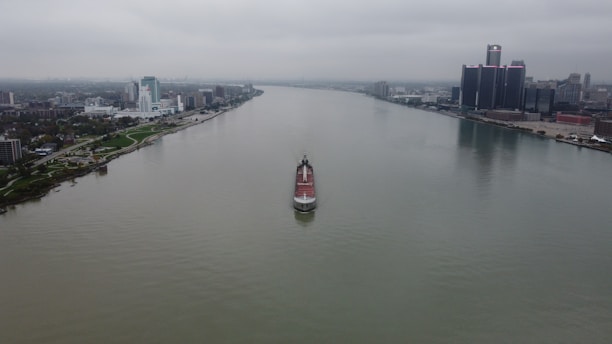 A large ship is cruising down a wide river with urban cityscapes on both sides. The skyline on the right is dominated by tall skyscrapers, while the left has smaller buildings and more greenery. The sky is overcast, creating a moody atmosphere.