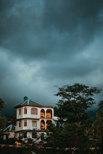 A multi-story house with arched windows and a sloped roof stands amidst lush greenery. The building is illuminated by warm, golden light against the backdrop of a dark, cloudy sky, suggesting an impending storm or late evening time.