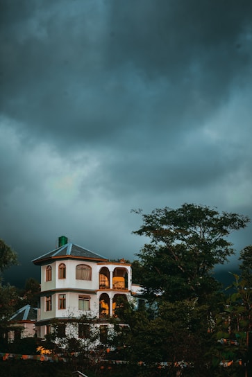 A multi-story house with arched windows and a sloped roof stands amidst lush greenery. The building is illuminated by warm, golden light against the backdrop of a dark, cloudy sky, suggesting an impending storm or late evening time.