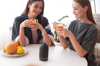 Two women sit at a table enjoying drinks from glass cups. A plate with citrus fruits, including oranges and lemons, is on the table. In the center of the table, there is a black aroma diffuser emitting steam. A plant in the background adds a touch of greenery to the setting.