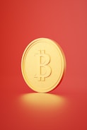 A gold-colored coin with the Bitcoin symbol embossed on it is displayed against a plain red background. The coin appears to be standing upright on its bottom edge, casting a soft shadow on the surface beneath it.