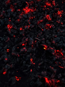 Dark charcoal pieces glow with vivid red and orange embers, creating a striking contrast against the surrounding blackened ash. The overall appearance suggests smoldering coals emitting warmth and light.