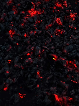 Dark charcoal pieces glow with vivid red and orange embers, creating a striking contrast against the surrounding blackened ash. The overall appearance suggests smoldering coals emitting warmth and light.