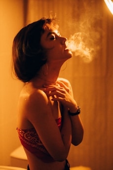 A person stands indoors with eyes closed, exhaling smoke against a dimly lit background. The lighting creates a warm, soft atmosphere, emphasizing the contours of their face and body. They have short hair and wear hoop earrings, a red bandana-style top, and a black wristwatch.