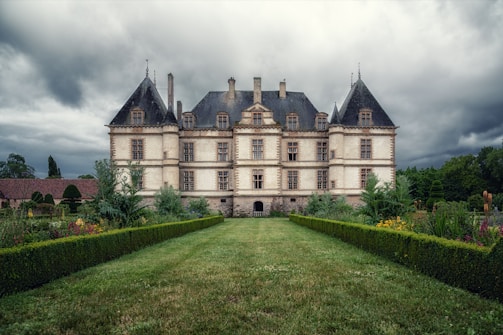 An imposing chateau with a symmetrical design stands amidst lush gardens. The building features a traditional French architectural style with tall, pointed roofs and numerous windows. The surrounding garden is neatly manicured with vibrant flowers and hedges, leading to the entrance of the chateau. Overhead, the sky is overcast, creating a dramatic contrast between the building and the clouds.