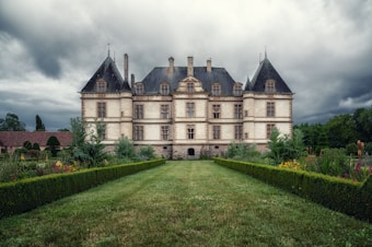 An imposing chateau with a symmetrical design stands amidst lush gardens. The building features a traditional French architectural style with tall, pointed roofs and numerous windows. The surrounding garden is neatly manicured with vibrant flowers and hedges, leading to the entrance of the chateau. Overhead, the sky is overcast, creating a dramatic contrast between the building and the clouds.