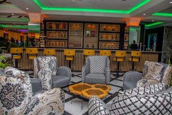 A modern bar interior with a stylish seating arrangement comprising patterned armchairs surrounding a geometric wooden table. The bar area in the background features a variety of bottles arranged on shelves with dramatic lighting. Bar stools are neatly lined up in front of the bar, and the ceiling is accented with green lighting, adding a vibrant yet relaxed atmosphere.