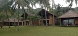 A large, rustic villa complex surrounded by lush greenery and tall palm trees. It features traditional thatched roofs and wooden structures, set against a backdrop of forested hills.