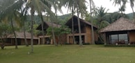A large, rustic villa complex surrounded by lush greenery and tall palm trees. It features traditional thatched roofs and wooden structures, set against a backdrop of forested hills.