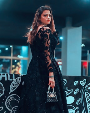 A woman in a long, elegant black lace dress stands indoors, posing with a small black handbag adorned with pearls. Her hair is styled in loose waves, and she looks over her shoulder with a confident expression. The background features abstract black and white art on a wall, which includes patterns and shapes like leaves and hearts. The setting is dimly lit with blurred lights in the distance, adding to the moody atmosphere.