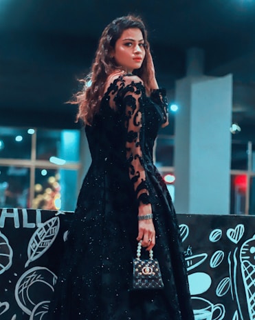 A woman in a long, elegant black lace dress stands indoors, posing with a small black handbag adorned with pearls. Her hair is styled in loose waves, and she looks over her shoulder with a confident expression. The background features abstract black and white art on a wall, which includes patterns and shapes like leaves and hearts. The setting is dimly lit with blurred lights in the distance, adding to the moody atmosphere.