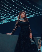 A woman dressed elegantly in a black lace gown adorned with intricate patterns stands in front of a backdrop of cascading string lights. She holds a small, embellished handbag and wears sparkling jewelry, including a choker and earrings. The ambiance suggests a night event with a sophisticated and glamorous atmosphere.