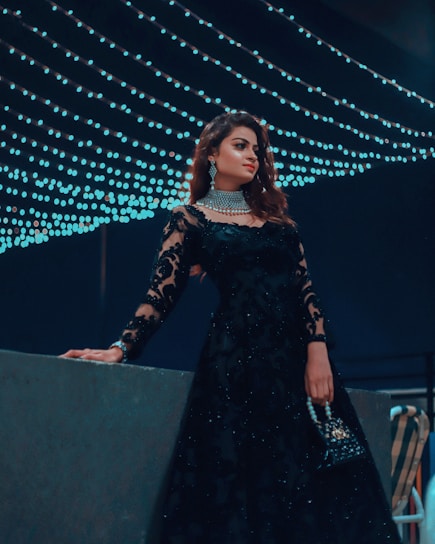 A woman dressed elegantly in a black lace gown adorned with intricate patterns stands in front of a backdrop of cascading string lights. She holds a small, embellished handbag and wears sparkling jewelry, including a choker and earrings. The ambiance suggests a night event with a sophisticated and glamorous atmosphere.