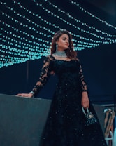 A woman dressed elegantly in a black lace gown adorned with intricate patterns stands in front of a backdrop of cascading string lights. She holds a small, embellished handbag and wears sparkling jewelry, including a choker and earrings. The ambiance suggests a night event with a sophisticated and glamorous atmosphere.